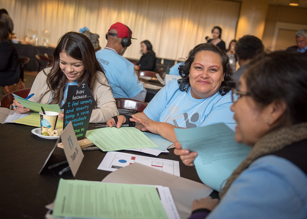 A photo of three women at a circular table, they are all different ages and ethnicities. The woman in the middle is looking at the camera and she is confident and happy. The group are sharing papers to read and talking to each other and the rest of their group.