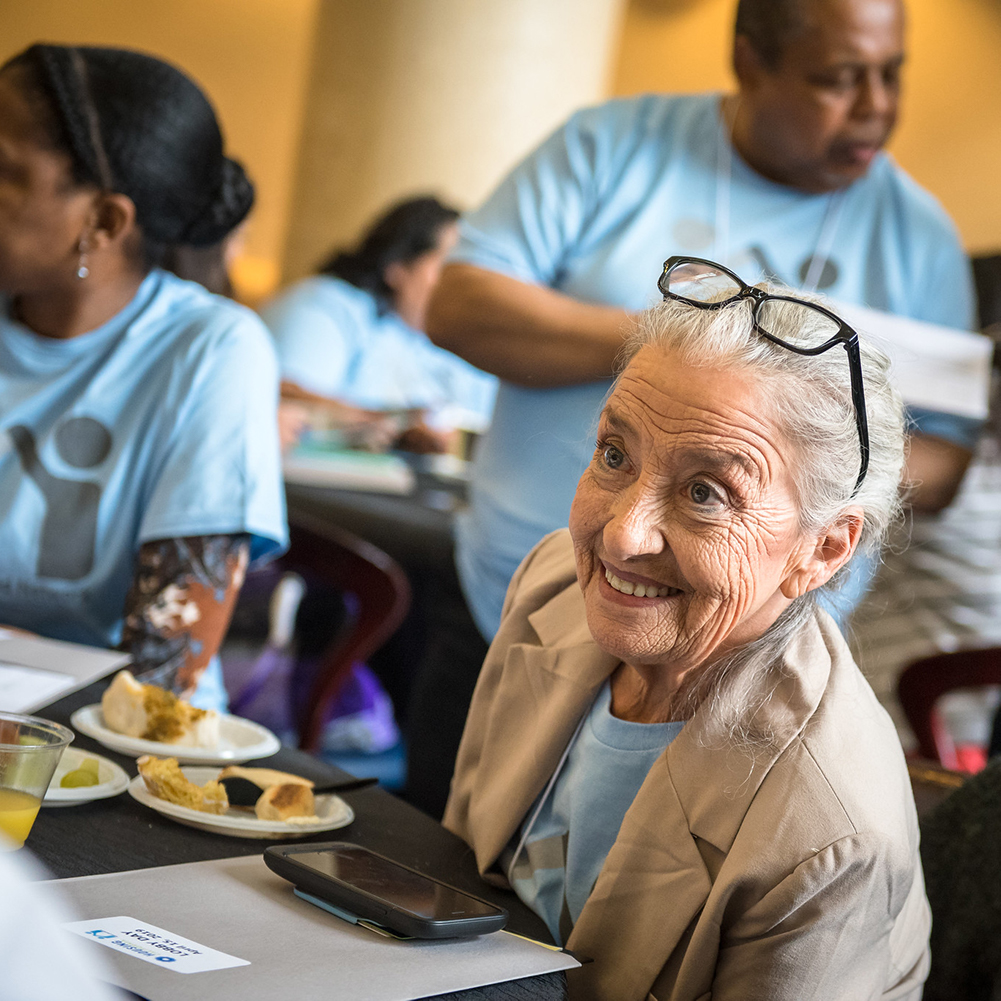 Close-up photo of an elderly white woman in a light blue t-shirt sitting at a table and talking with another Lobby Day attendee off camera. She has white hair, a big smile, and glasses perched on her head.