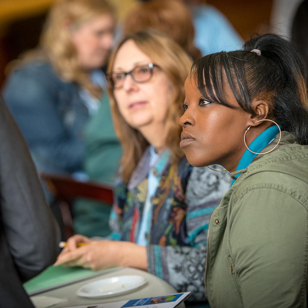 Close-up photo of a young black woman sitting at a table, she is in a room with many other people and listening intently to a speaker. 