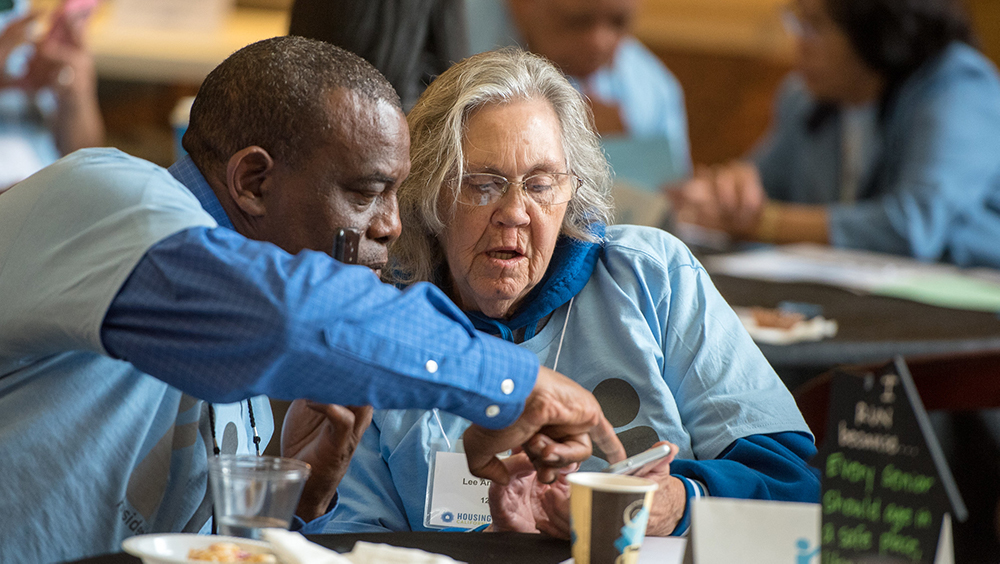 Two seniors, an African American man and a caucasian woman in glasses, study something on the woman's phone. They are wearing light-blue t-shirts and are attending Lobby Days.