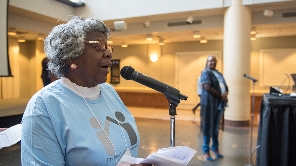 An African American woman with curly gray hair, red-rimmed glasses, and a light blue t-shirt, stands at a microphone. She holds a paper in her hand, her head is held high and she is talking to people off camera to the right of the image.