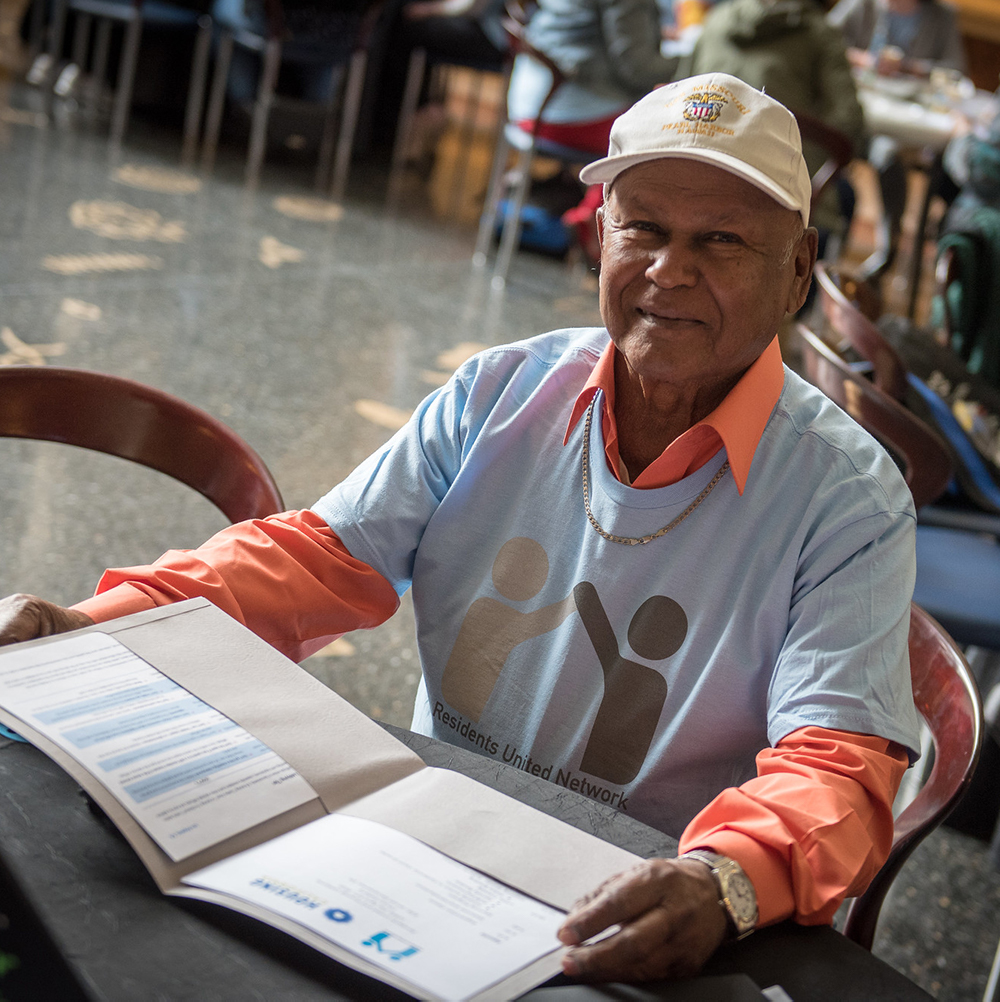 Close-up photo of a senior man in a ball-cap and a light blue t-shirt, sitting at a circular table reading a folder with information about Residents United Network. He is looking at the camera with a slight smile, and is surrounded by lots of other people at tables too. It is Lobby Day for RUN leaders.