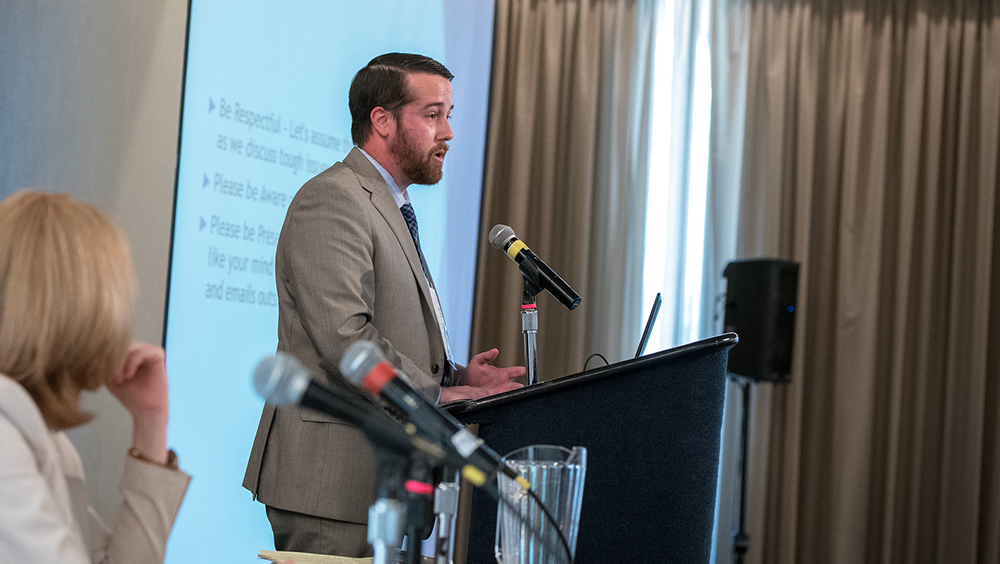 Side profile of Chris Martin, from Housing California, standing at a podium with a microphone and talking. In the background is a projection screen with a text slide showing.