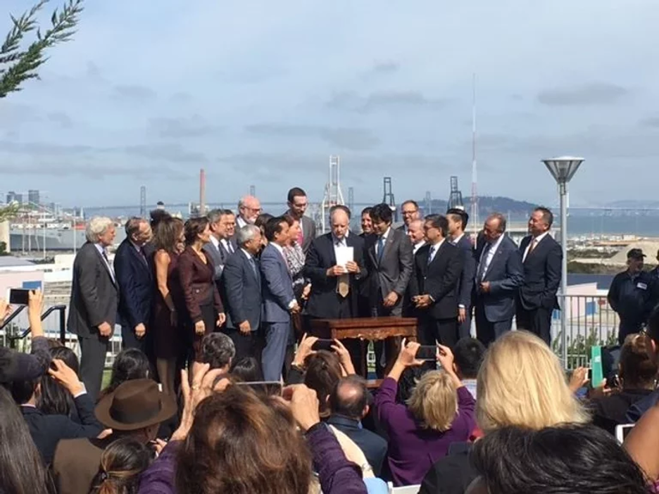 A group of Housing California supporters stands on a raised platform in San Francisco by the water, in front of a crowd. Governor Jerry Brown is on the center of the group, and Housing California Executive Director, Lisa Hershey, is also on the stage. The crowd are taking photographs with their smartphones.