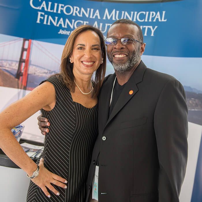 At the Housing California Annual Conference, Executive Director Lisa Hershey smiles for the camera with her arm around a smiling black gentleman in glasses and a suit. They are standing at the supporter booths, and the banner behind them says California Municipal Finance Authority.