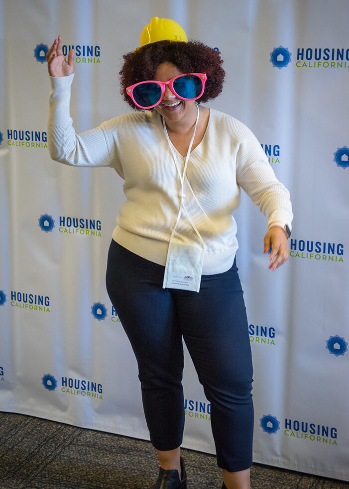 A woman with curly hair strikes a goofy pose at the photo booth. She has one arm in the air and looks like she is dancing. She is wearing a tiny yellow hard-hat and very large pink-rimmed clown glasses. She is having a great time at the Annual Conference!