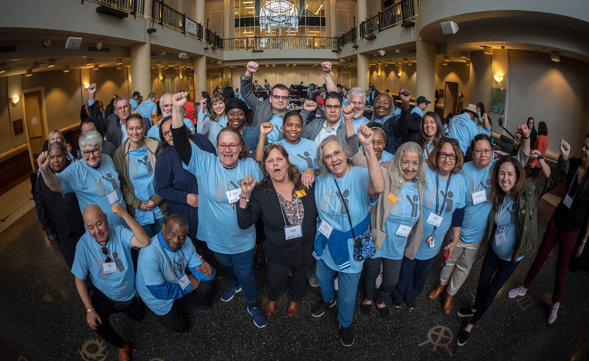 A group of around 30 people are standing inside a large well-lit hall. They are looking at the camera and cheering. Everyone is wearing a light blue t-shirt with Residents United Network on the front. The people are all different ages and ethnicities, and they look accomplished, and powerful. 