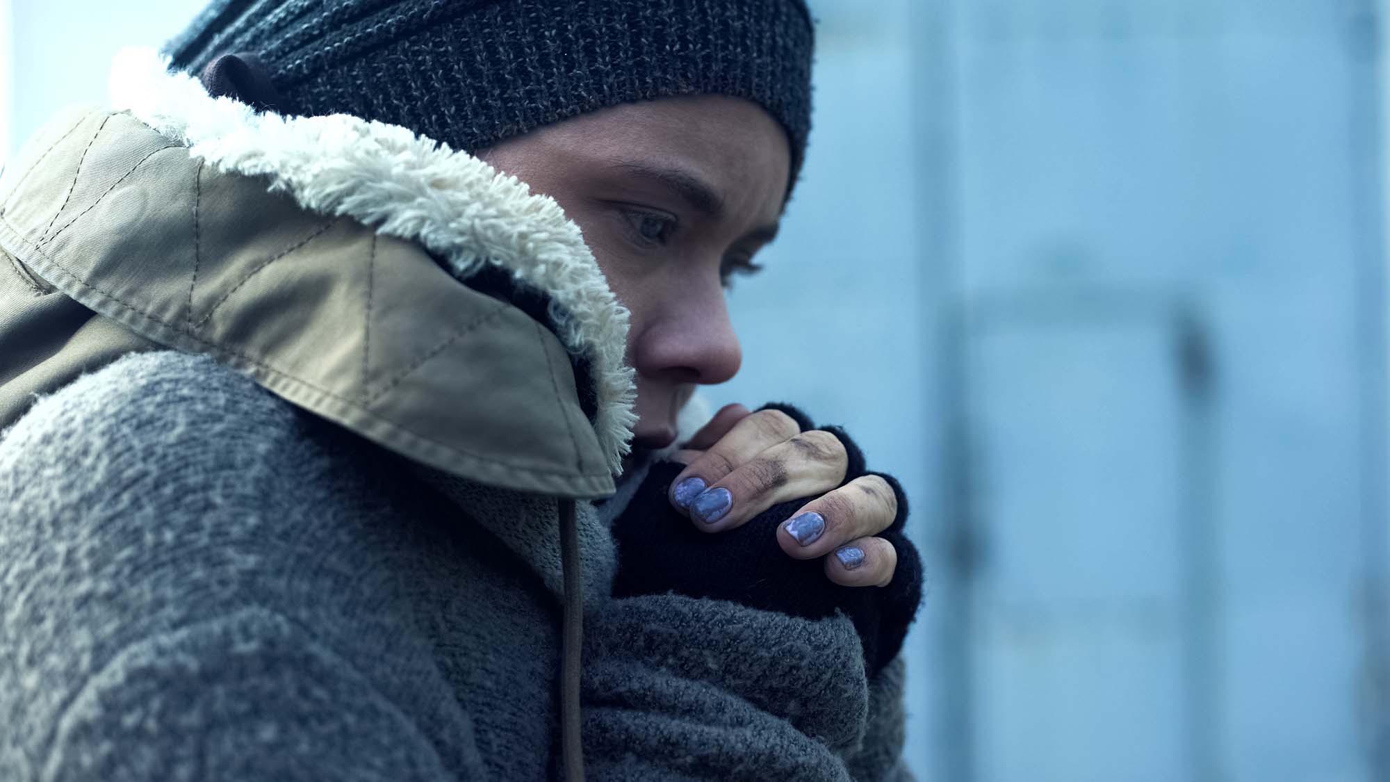 A woman in a knitted hat, fingerless gloves, chipped nail polish, and a large jacket with a furry hood comforts herself in the cold. Her clasped hands are at her chin, and she stares slightly downwards. She appears to be lonely.