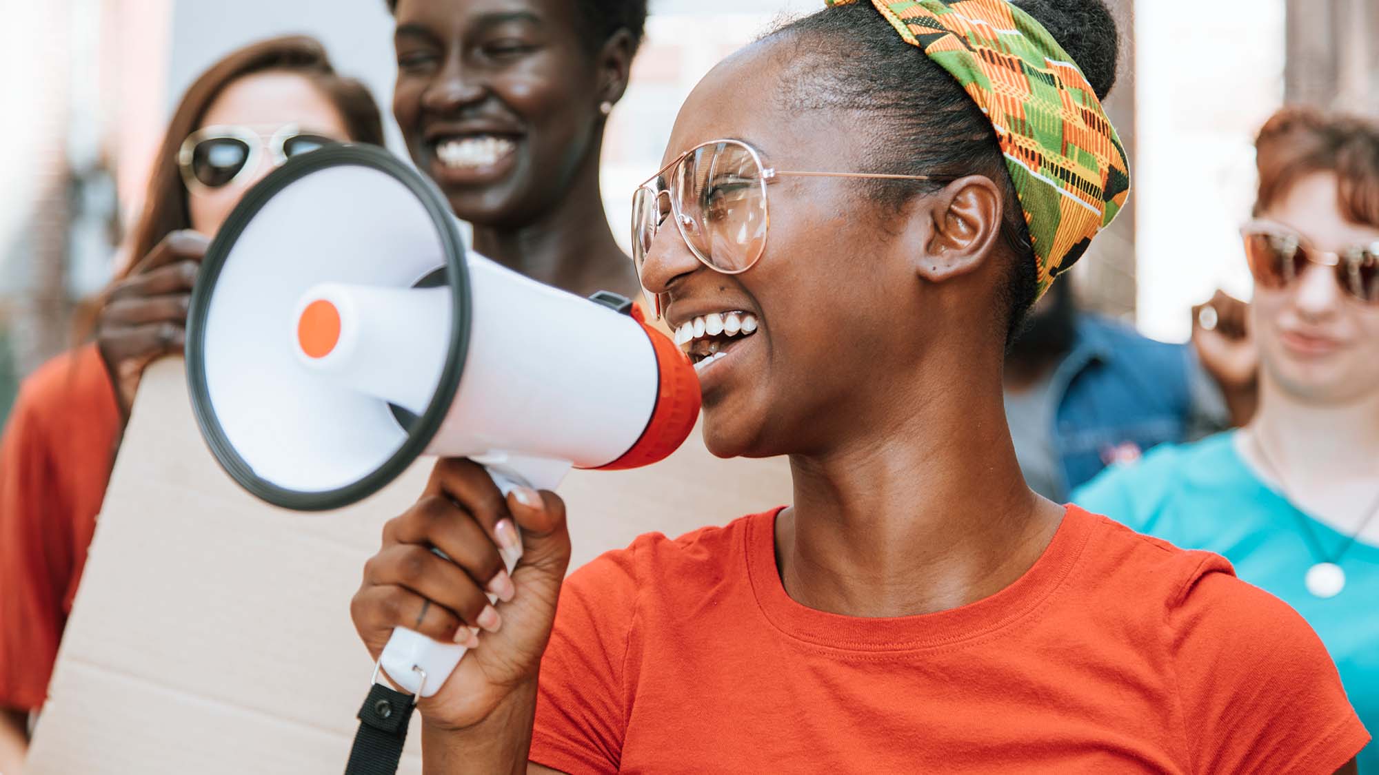 Young African American woman leads a group of people in making their voices heard. They are walking on the street and she has a mega-phone up to her mouth. She is smiling and positive in a colorful headband and red t-shirt.