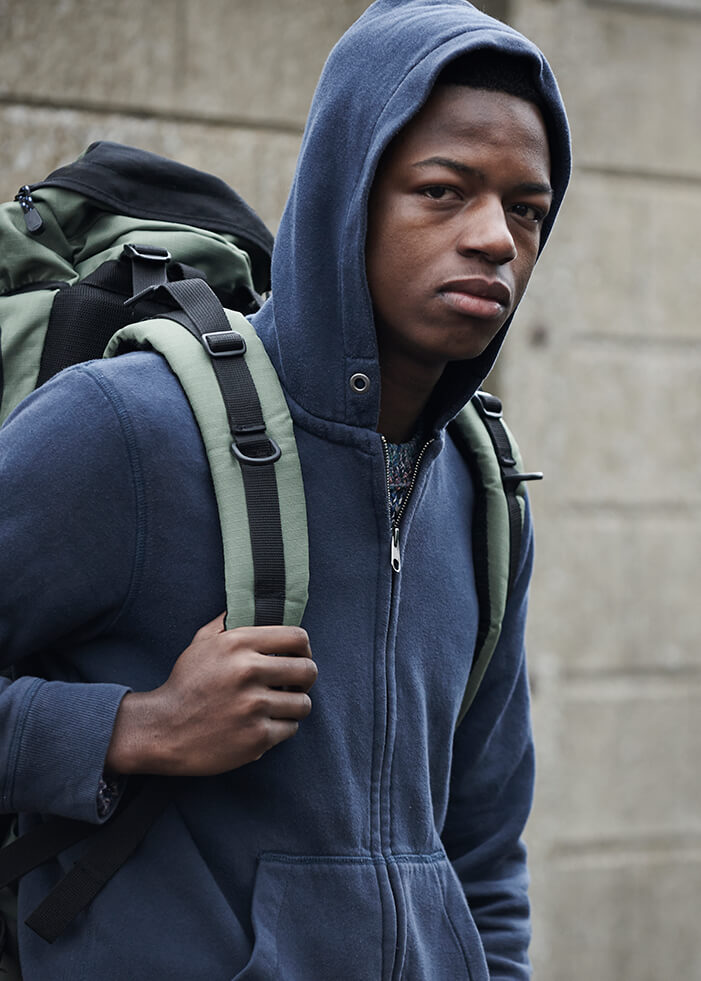 An African American teenage boy stands in front of a large gray wall, his right arm is bent at the elbow and he holds onto the strap of his green backpack. He wears a dark blue hooded sweater. The boy is facing to the right, but his head is looking at the camera, and he looks sad. The concept is homeless youth in California are disproportionately people of color.