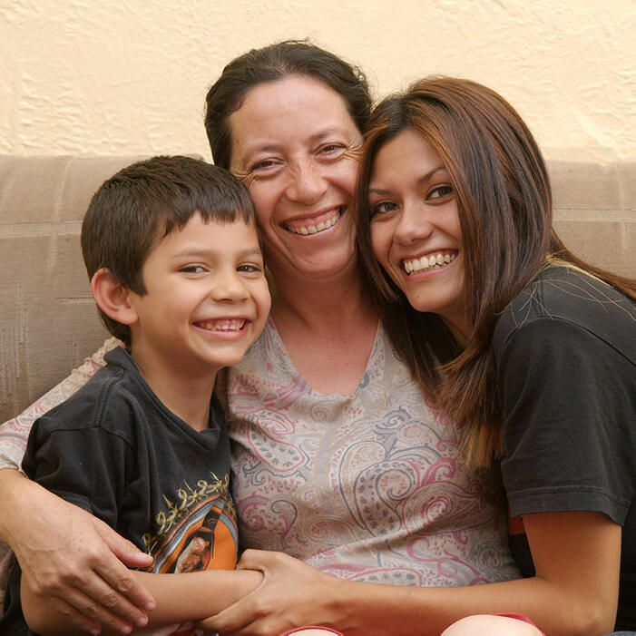 A photo of Maria and her teenage daughter and young son sitting on the couch in their rental home. The children sit on each side of Maria, and she wraps her arms around them. Everyone is smiling at the camera.