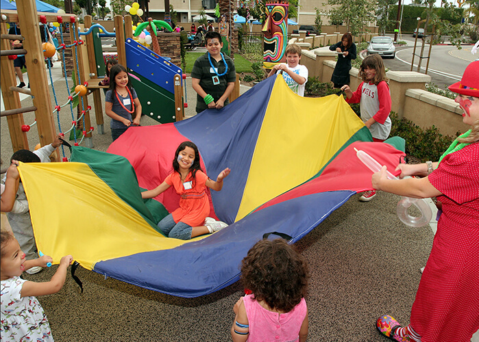 A photo of children playing with a multi-colored parachute in a park connected to affordable apartment homes. They are holding up the parachute while one girl sits in the middle, laughing. The children are very active, smiling and having fun; some of the children have painted faces. A woman dressed as a clown with big shoes and a red bowler hat provides supervision, and laughs with the children.