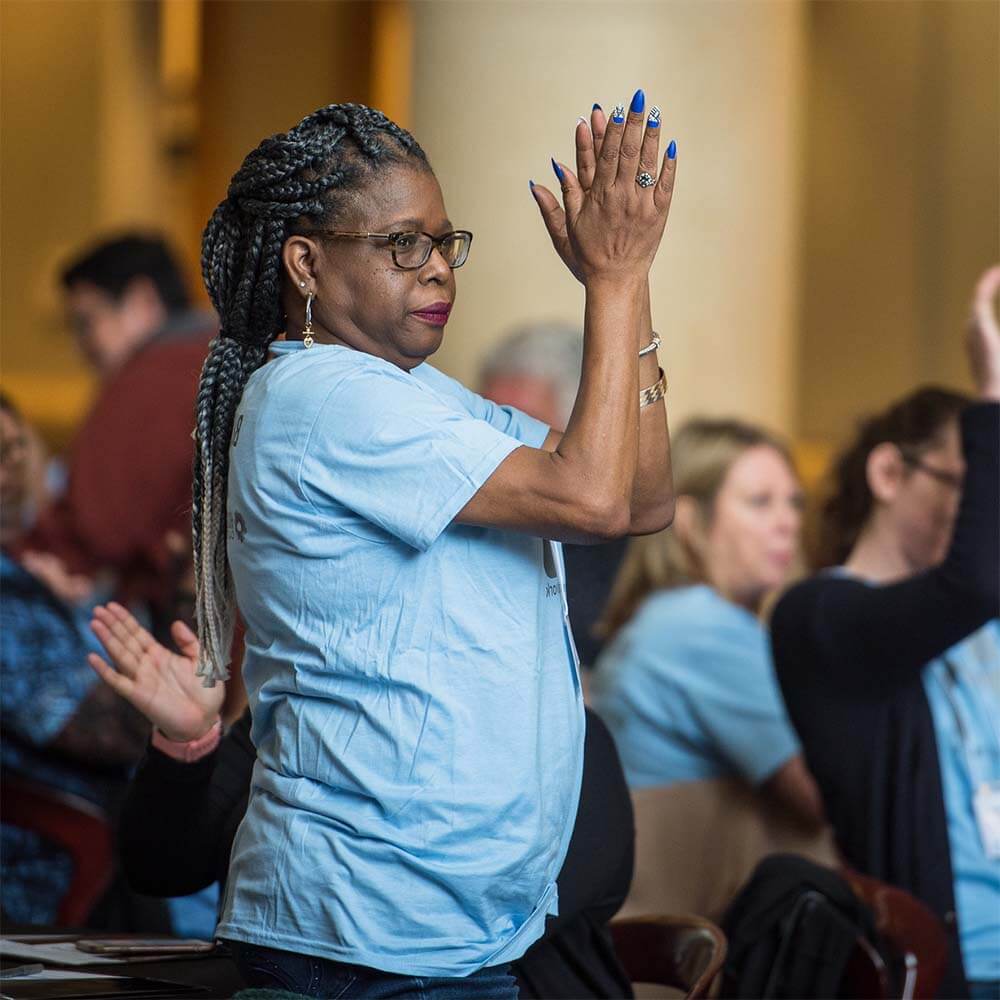 An African American woman with long brown braided hair, wearing brown-rimmed glasses and a light blue t-shirt, stands facing to the right, her hands are raised to the height of her forehead and she is clapping. Her long nails are painted blue to match the Residents United Network brand. She looks confident and proud.