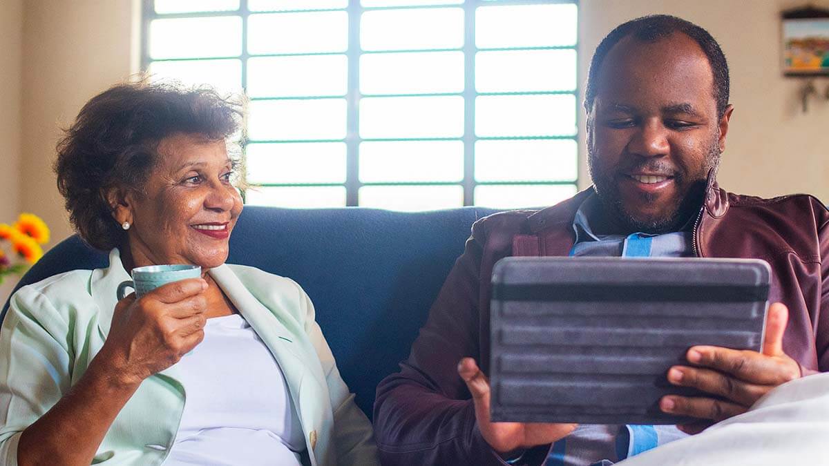 Portrait of a senior mother and her adult son sitting on the couch in front of a bright window. The mother sits on the left, smiling and looking at her son. She is holding a mug. The son is smiling and looking at the digital device he is holding in both hands. They look happy and content in each other's company.