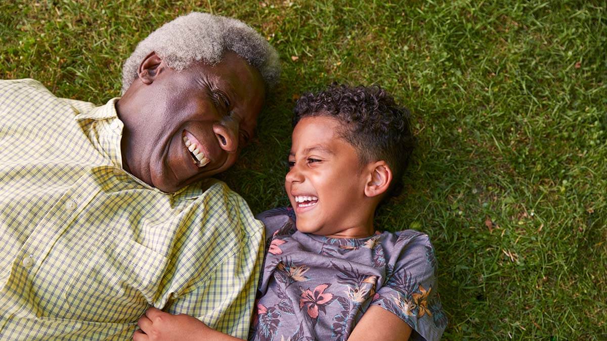 A Grandpa and his young grandson, dressed in bright shirts, lay on their backs on the grass, looking at each other, and laughing.