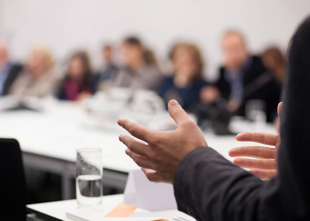 A meeting or presentation in progress. The background is blurred and in it are people who are seated, wearing business attire. In the foreground is a partial view of a person talking to the group, with the person's hands visible and gesturing to the group.