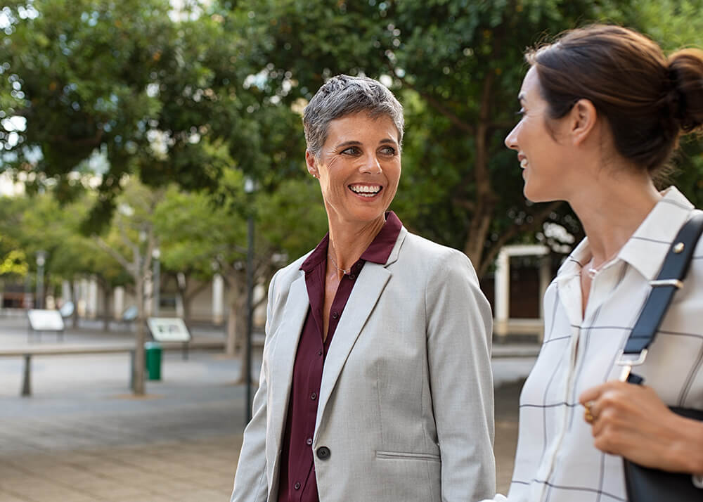 Two women wearing office attire walking side-by-side, smiling at each other. The woman on the left has short gray hair, and is wearing a burgundy dress shirt with a gray jacket. The woman on the right is wearing a white dress shirt with dark lines, and carrying a dark purse on her shoulder.