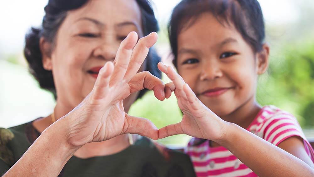 A grandmother and grand-daughter, both of Asian/Pacific Island ethnicity, are shoulder-to-shoulder and facing the camera. Their hands come together in the center, thumbs and forefingers touching and forming a heart shape.