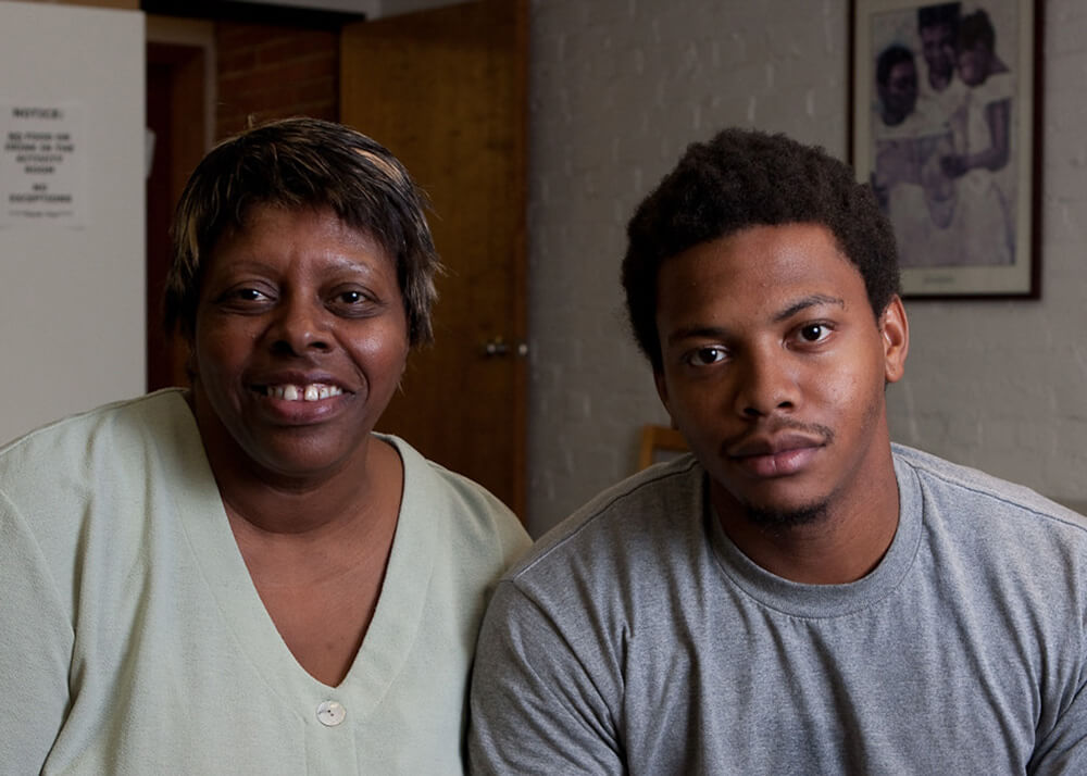 A photo of a mother on the left, and her adult son on the right, they are sitting and looking at the camera. The woman's name is Wenda, and she has been a hard worker and homeowner for most of her life. A series of unexpected medical conditions and bills resulted in Wenda losing her home, and Wenda and her son found themselves on the streets with nowhere to turn. This image was taken the day Wenda received news that she could get temporary accommodation while she looked for a new place to call home. Wenda is smiling, and her son looks relieved.