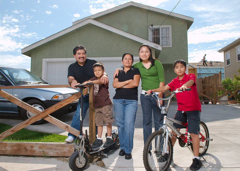 Eufemio and Leticia, Michelle, Alex and Jesse, stand outside their new house in Greater Los Angeles. Alex sits on a bike and his younger brother stands on a scooter. Sandra has her arm around Mom's shoulder. There is a car in the driveway, and in the background two people are standing on roof rafters, they are building a new house. This home is one of several built by homeowners and volunteers with Habitat for Humanity.