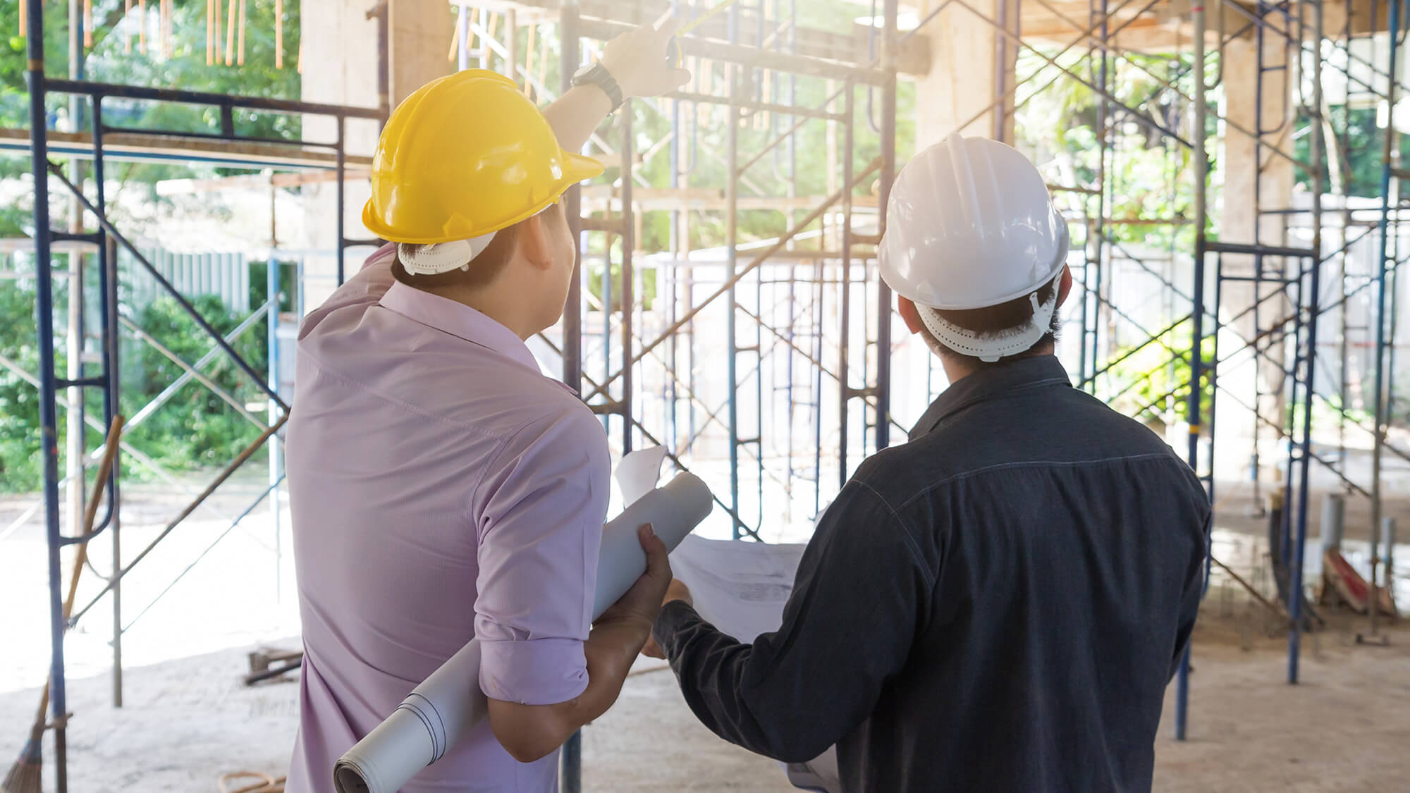 Two people stand on concrete inside scaffolding at a building site. The site is very open and sunny. The people both wear hard-hats and collared shirts. The person on the left, holding rolled plans under their arm, points to the upper levels of the scaffolding. The person on the right holds a plan out in front but looks toward the higher levels, as well.