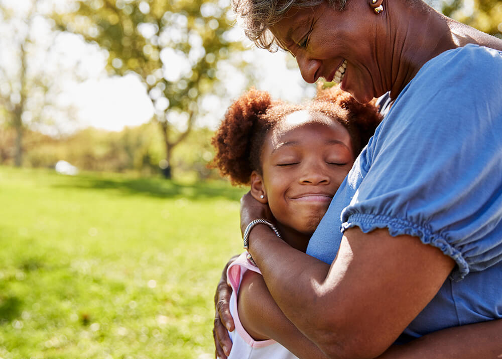 A close-up photo of a grandmother hugging her young granddaughter in the park. The young girl's head rests under her grandmother's chin, her eyes are closed and she is cherishing the moment, with her face toward the camera. The grandmother's face tilts down to the granddaughter, and she has a really big smile.