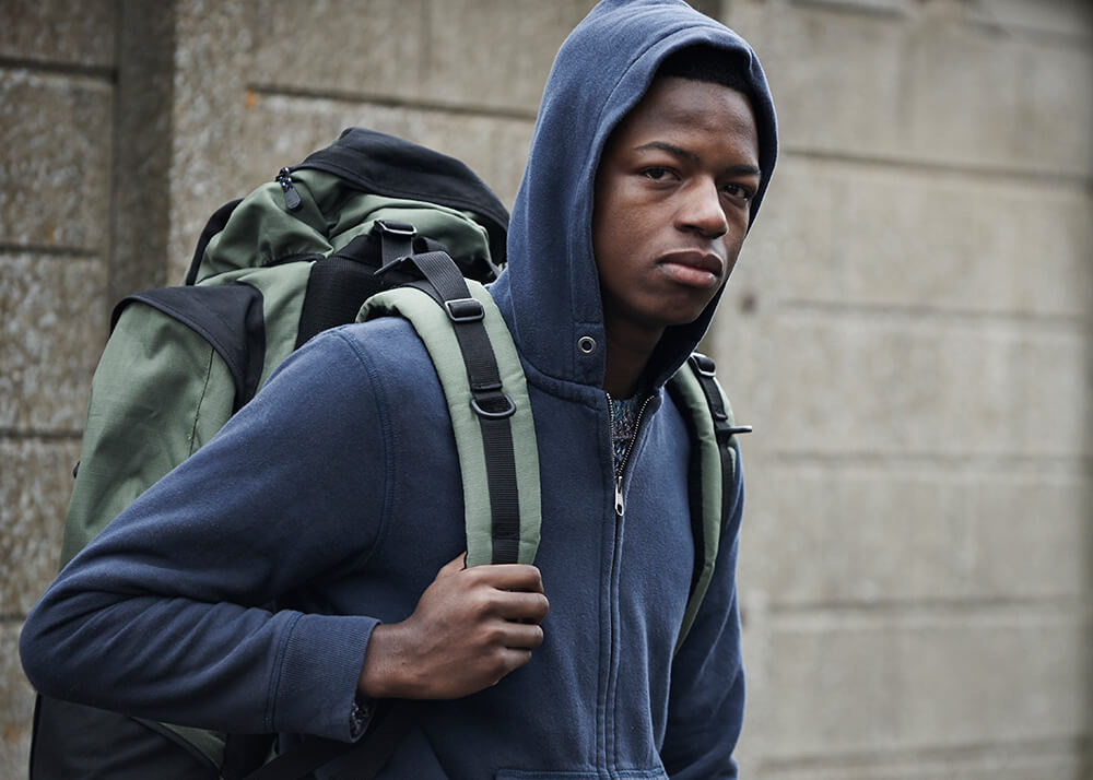 An African American teenage boy stands in front of a large gray wall, his right arm is bent at the elbow and he holds onto the strap of his green backpack. He wears a dark blue hooded sweater. The boy is facing to the right, but his head is looking at the camera, and he looks sad. The concept is homeless youth in California are disproportionately people of color.