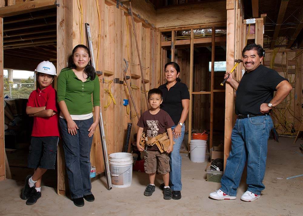 A Latino family stand inside of a partially constructed house. Mom, Dad and three children look at the camera. Dad stands on the right holding a hammer and is smiling. Two young boys wear a tool-belt and a hard-hat, they look confident. There are buckets and wires around them. 