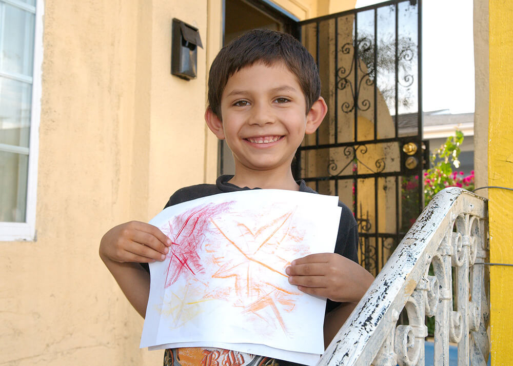 This is a photo of a boy named Guillermo. He is a kindergartner and 5 years old. Guillermo is growing up safe and secure with his family in a stable home, where he can do his homework and create art. He is pictured on the steps of his home, he has a huge smile, and he is holding his art in front of him for us to see. He has created leaf rubbings in red, orange, and yellow.