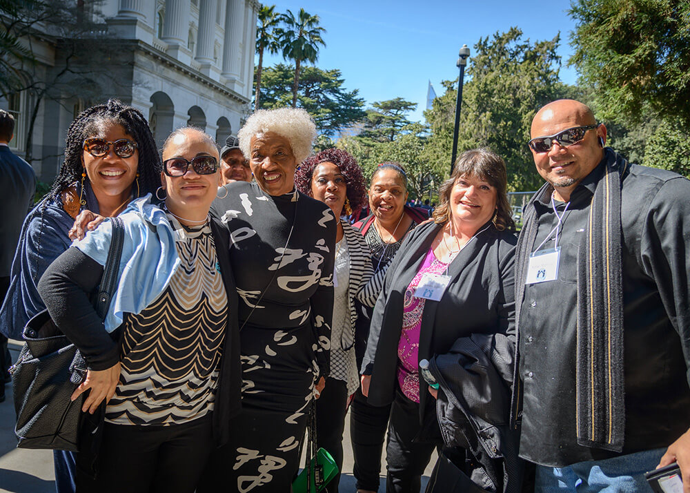 A small group of people stand outside of the Capitol in Sacramento, California. It's a sunny day, the sky is blue, they are wearing smart clothes, and they look very happy. The people are members of Residents United Network and they are attending the annual Lobby Day where they get to tell their stories to representatives.