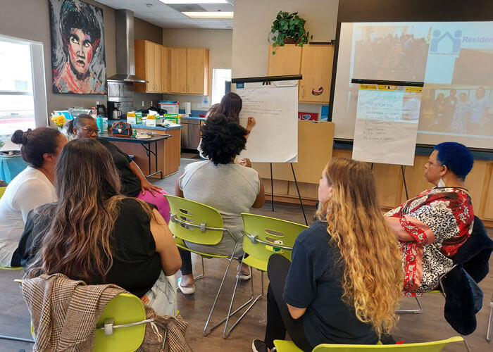 A small group of female affordable housing residents sitting together to discuss policy proposals, while a female with long brown hair stands in front of them, writing on a large easel with paper.