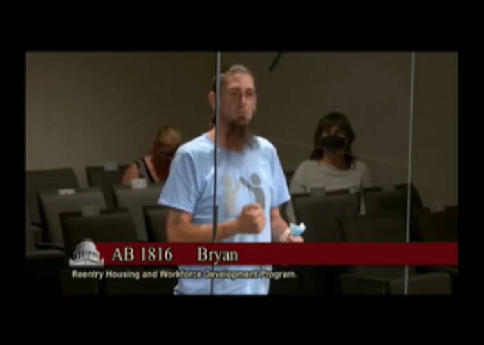 Video screenshot of a young male RUN leader with a brown chin beard and wearing a light blue RUN t-shirt, speaking to a group of people to advocate for Assembly Bill 1816.