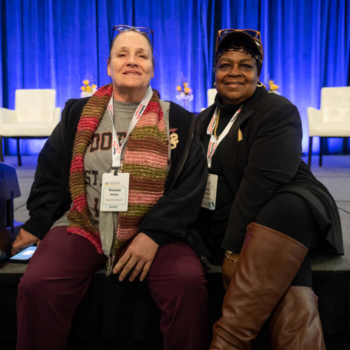 A caucasian woman on the left and african-american woman on the right sitting next to each other on the edge of a conference stage smiling towards the. camera