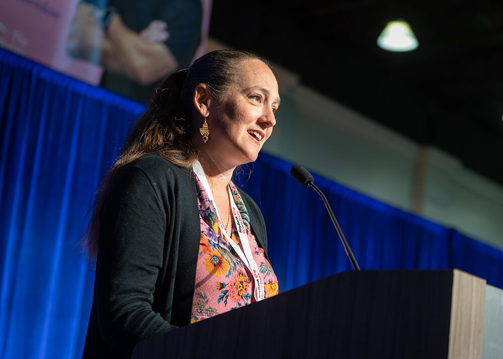 Housing California's Executive Director, Lisa Hershey, stands on a stage in front of a tall blue curtain. She wears a microphone attached to her dress collar. Her hands are animated as she talks energetically to the attendees of Lobby Days at the Capitol in Sacramento, California. She looks determined.